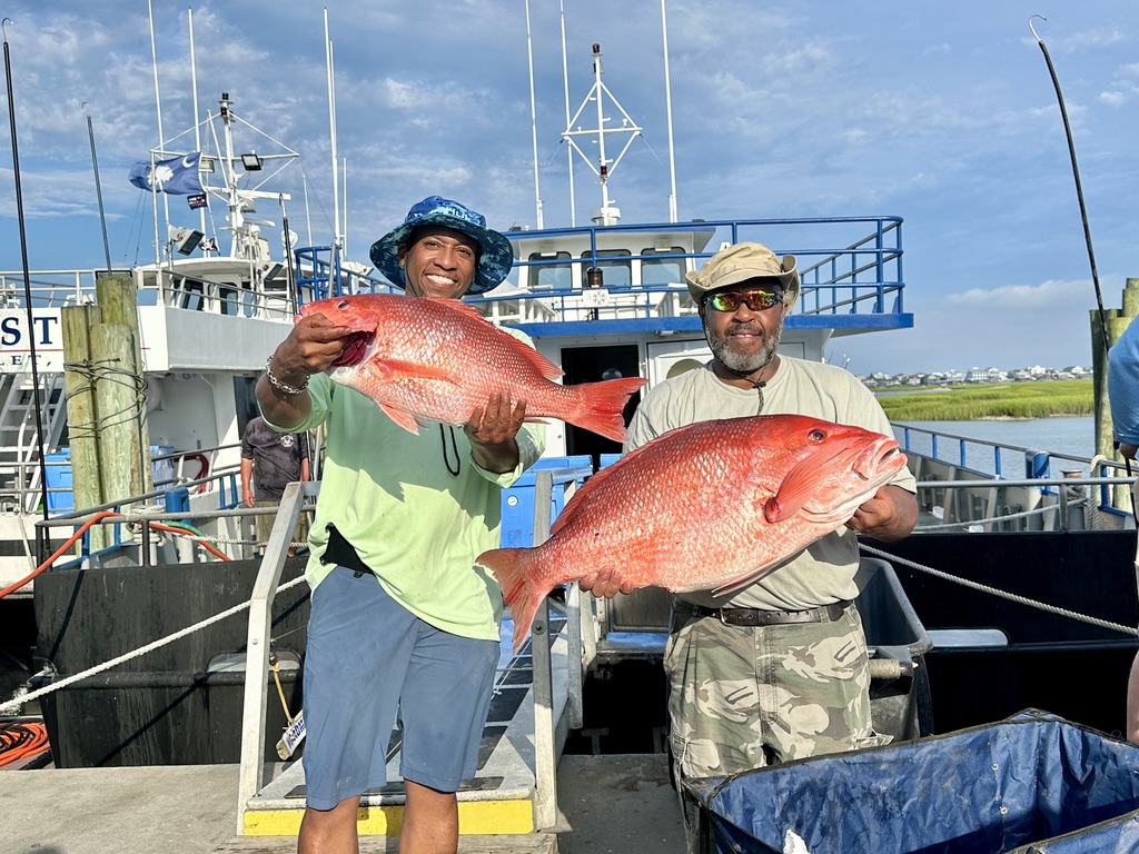Two men smiling, holding large red fish on a dock with boats in the background.