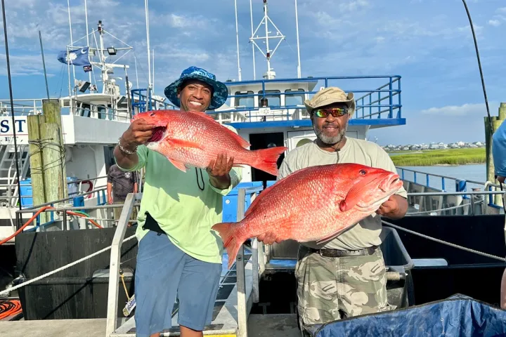 Two men smiling, holding large red fish on a dock with boats in the background.
