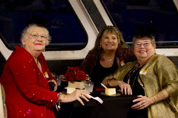 Three women in sparkly attire sitting at a table with cake and roses.