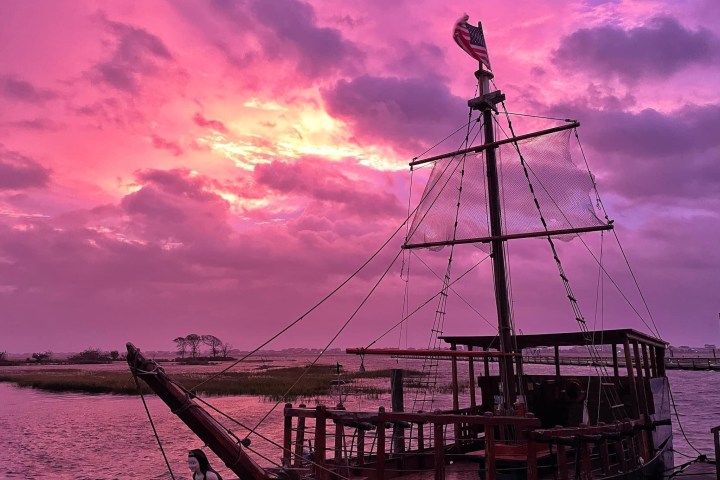 Old ship with sails against dramatic pink sunset over water.
