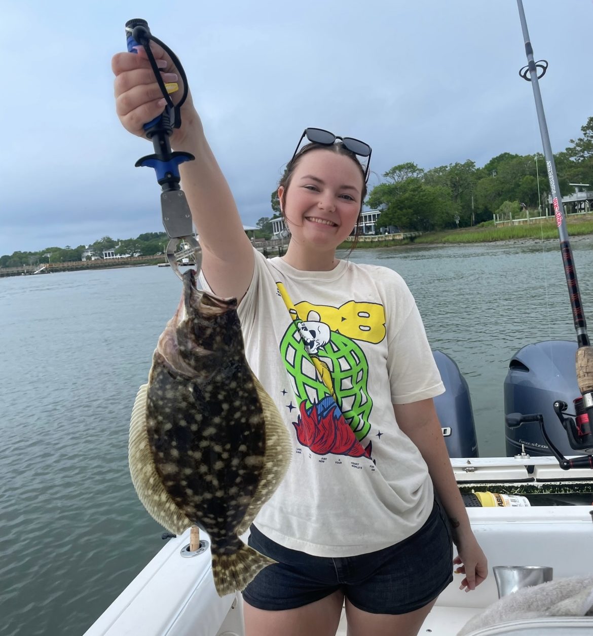 Person on boat in Murrells Inlet, SC holding a flounder fish with a fishing tool, smiling, wearing a graphic tee and shorts.