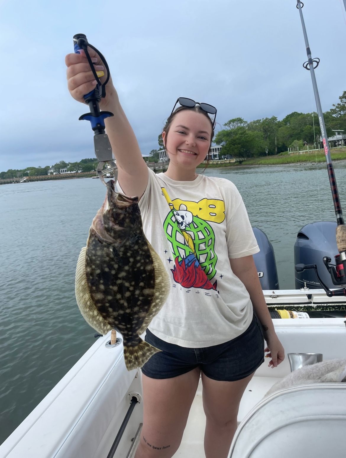 Screenshot Person on boat holding a fish with a fishing tool, smiling, wearing a graphic tee and shorts.