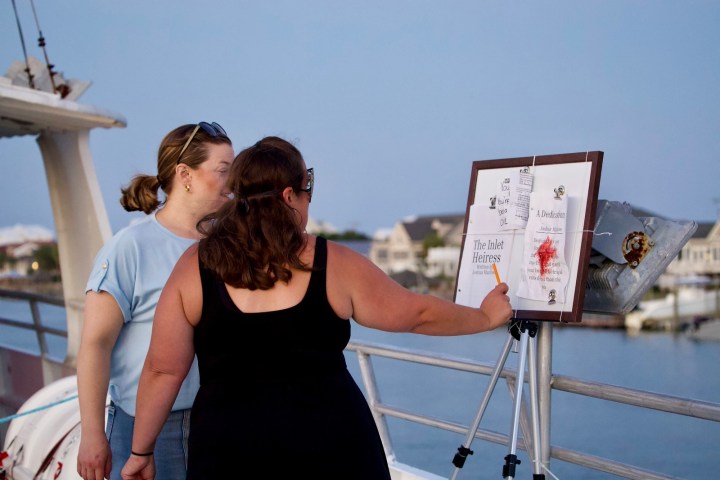 Two women examine a stand with papers titled 'The Inlet Heiress' on a boat deck.
