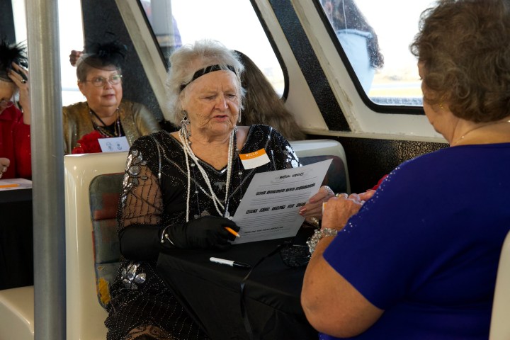 Elderly woman in costume reading paper across from another woman, on a boat or train.