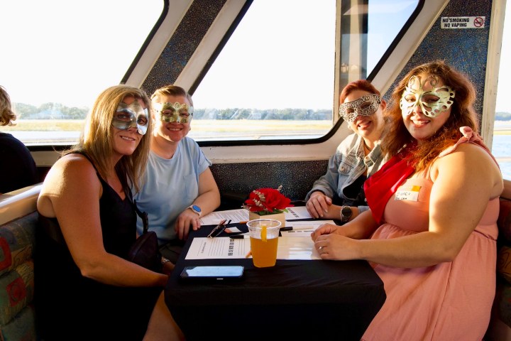 Four people wearing decorative masks seated at a table on a boat during the day.