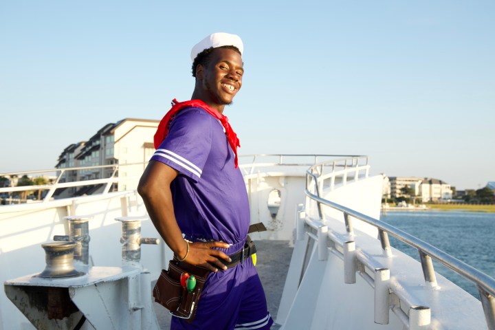 Person in sailor costume standing on a ship's deck, smiling broadly.
