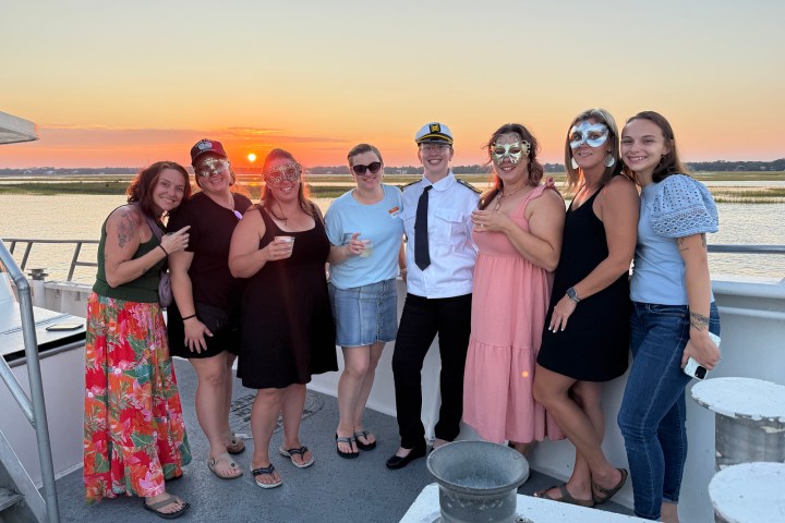 Group of people in festive attire on a boat at sunset, some wearing masks and one dressed as a captain.