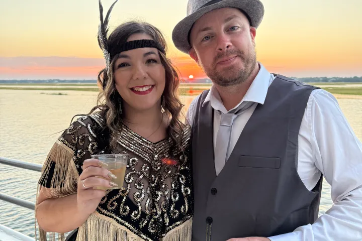 Couple dressed in 1920s attire smiling with drinks at sunset by the water.