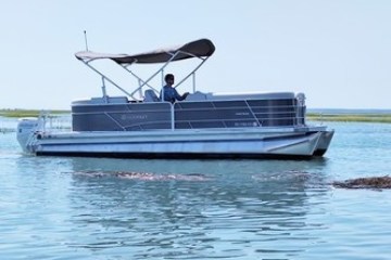 A pontoon boat with canopy and person navigating on calm water with grassy shoreline.