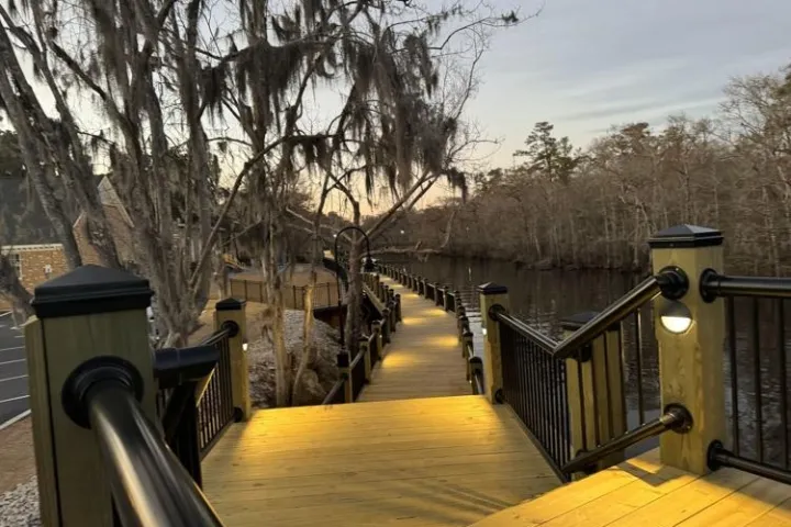 Well-lit wooden boardwalk with railings along a river, surrounded by trees at dusk.