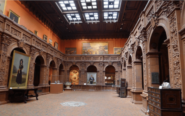 Ornate gallery with carved arches, paintings, and dim skylight.