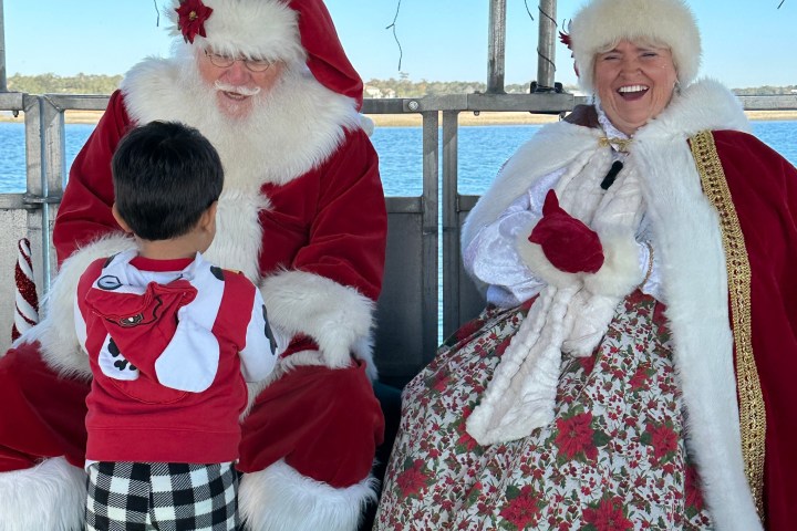 Child with Santa and Mrs. Claus on a boat, water in the background, smiling and festive.