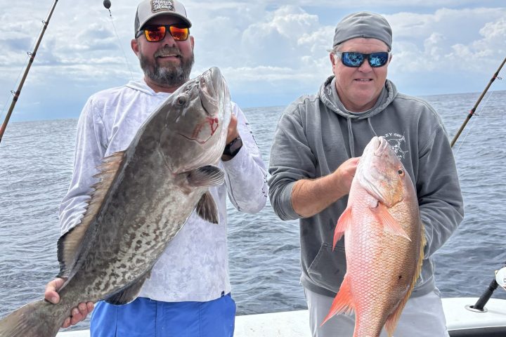 Two men on a boat holding large fish with fishing rods in the background.
