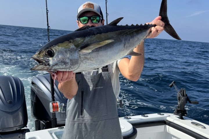 Person holding a large fish on a boat, wearing fishing gear, ocean in the background.