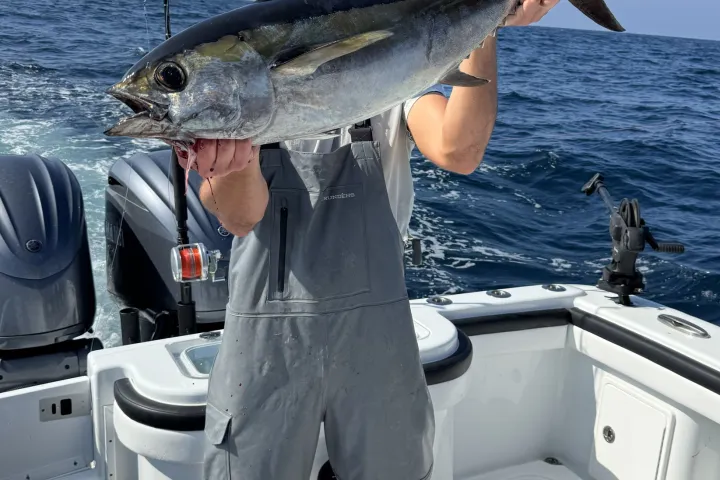 Person holding a large fish on a boat, wearing fishing gear, ocean in the background.