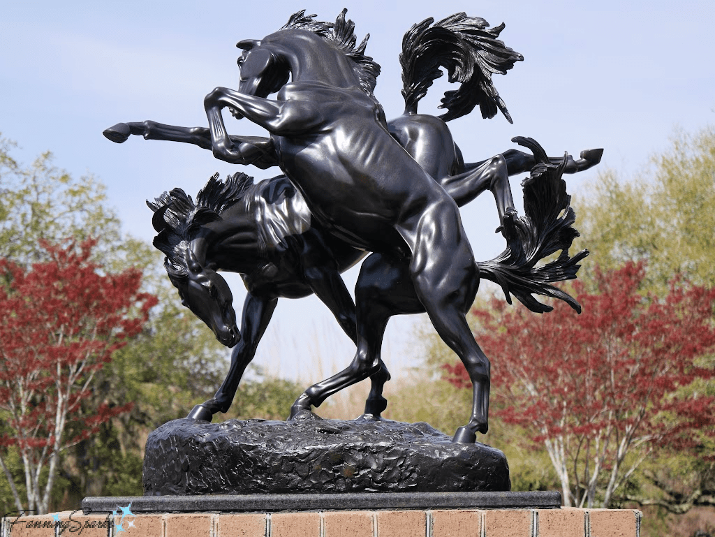 Bronze statue of two rearing horses on a stone base, with trees and sky in the background.