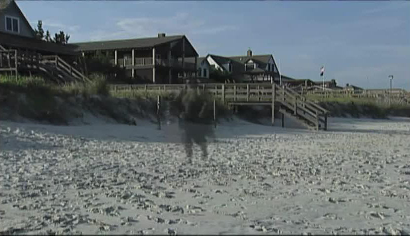 Shadowy figure on a sandy beach with houses and stairs in the background.