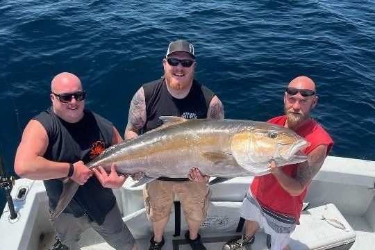 Three men on a boat holding a large fish over the ocean.