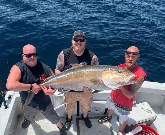 Three men on a boat holding a large fish over the ocean.