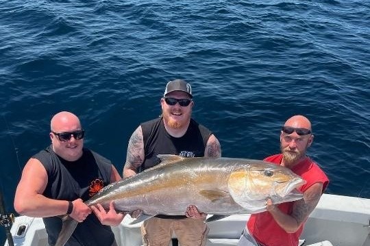 Three men on a boat holding a large fish over the ocean.