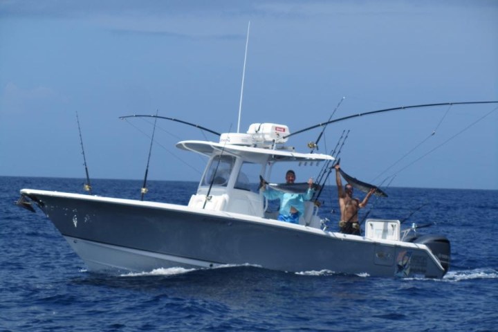 Two people on a boat holding fishing rods, sailing on blue ocean under a clear sky.