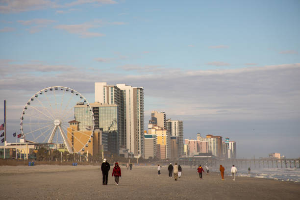 Beach scene with people walking, ferris wheel, and city skyline in the background.