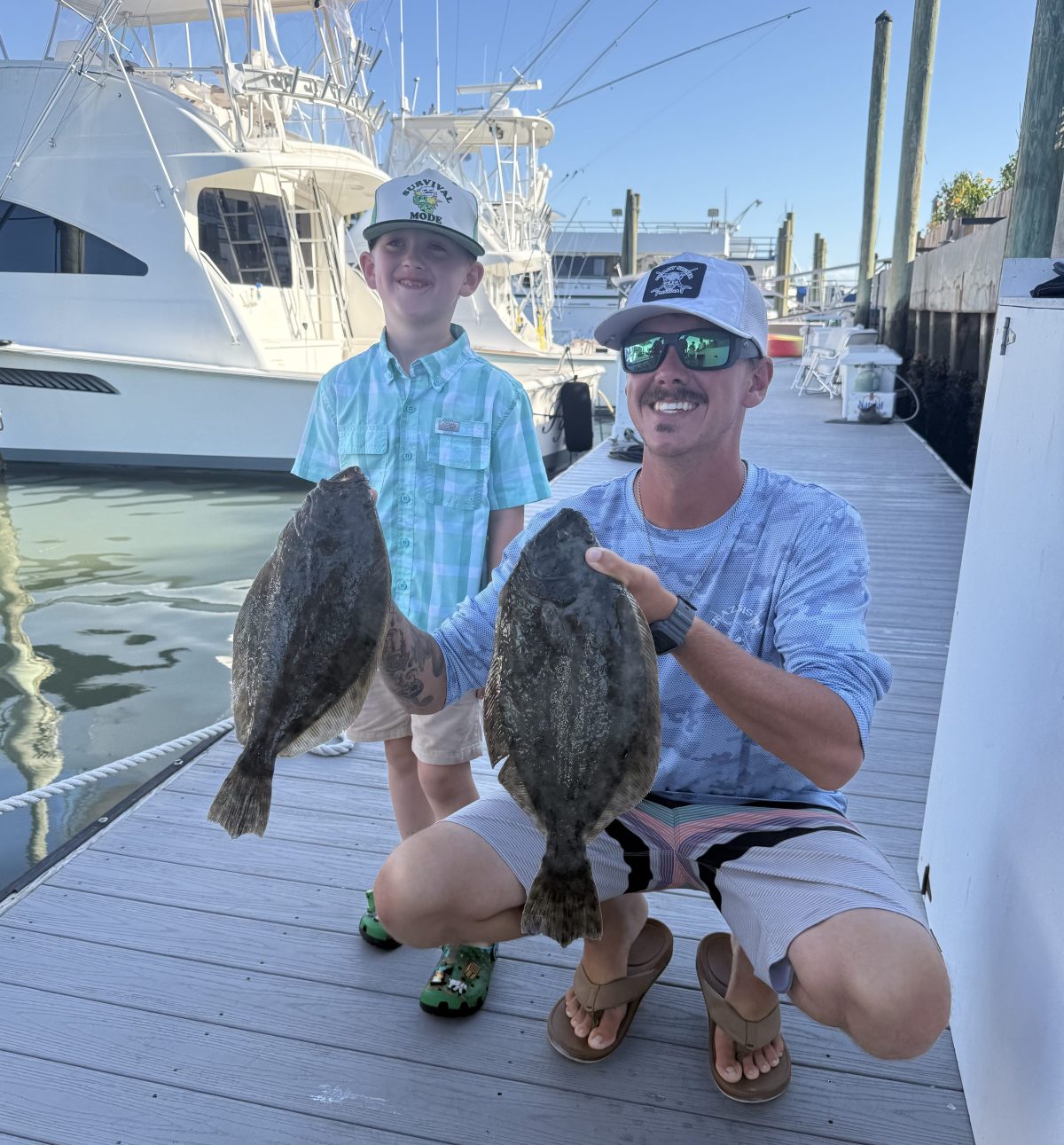 Man and child holding two flounder on a dock with boats in the background at Crazy Sister Marina in Murrells Inlet, SC.