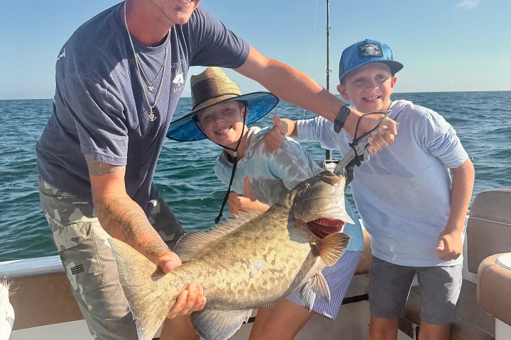 Three people on a boat holding a large fish, smiling with ocean in background.
