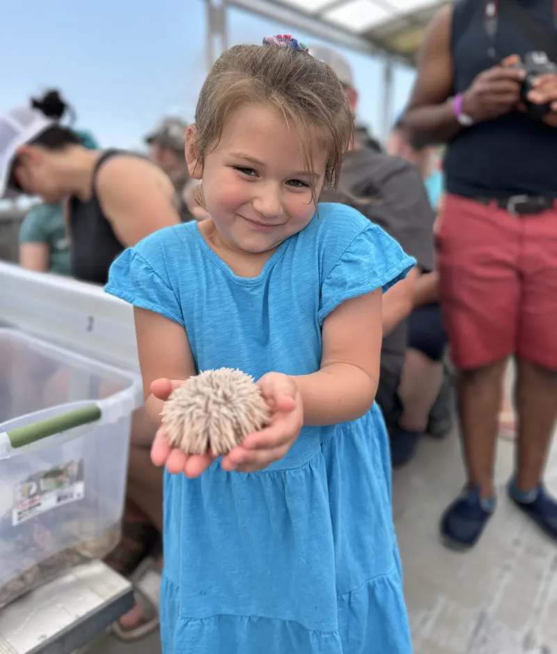 Smiling girl in blue dress holding a fluffy object, surrounded by people on a boat.