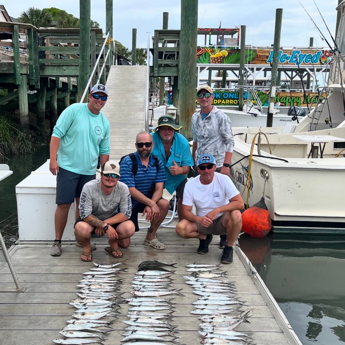 Group of six men posing on a dock with rows of fish, boats in the background.