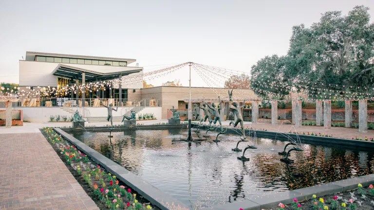 Modern building with glass walls, fountain sculptures, string lights, and a tree on a clear day.