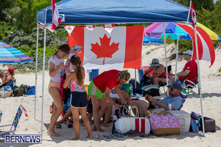 People relaxing under shade with Canadian flag on a sandy beach.