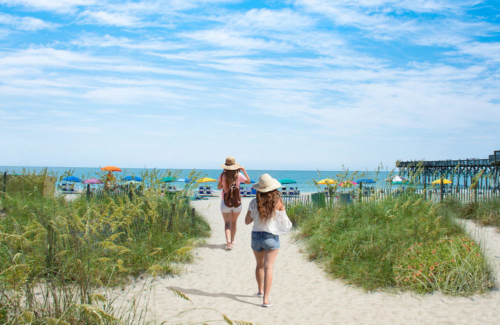 Two women walking on a sandy beach path towards the ocean under a blue sky.
