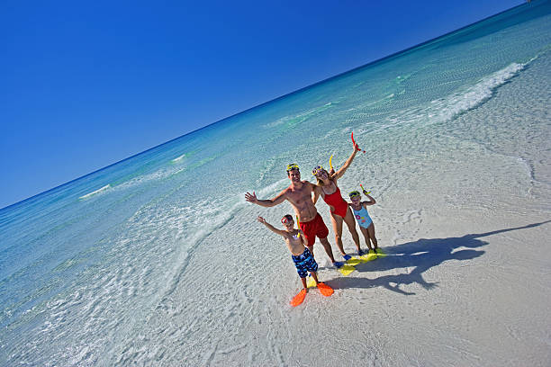 Family in swimwear with snorkels and flippers, standing happily on a sunny beach with clear blue water.