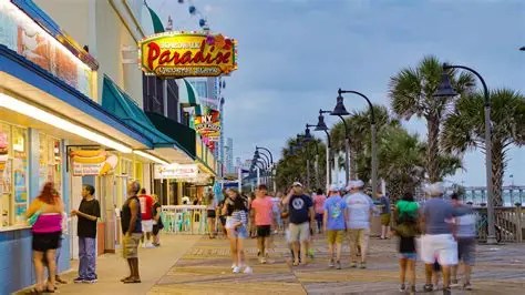 People walking on a bustling boardwalk lined with shops and palm trees at dusk.