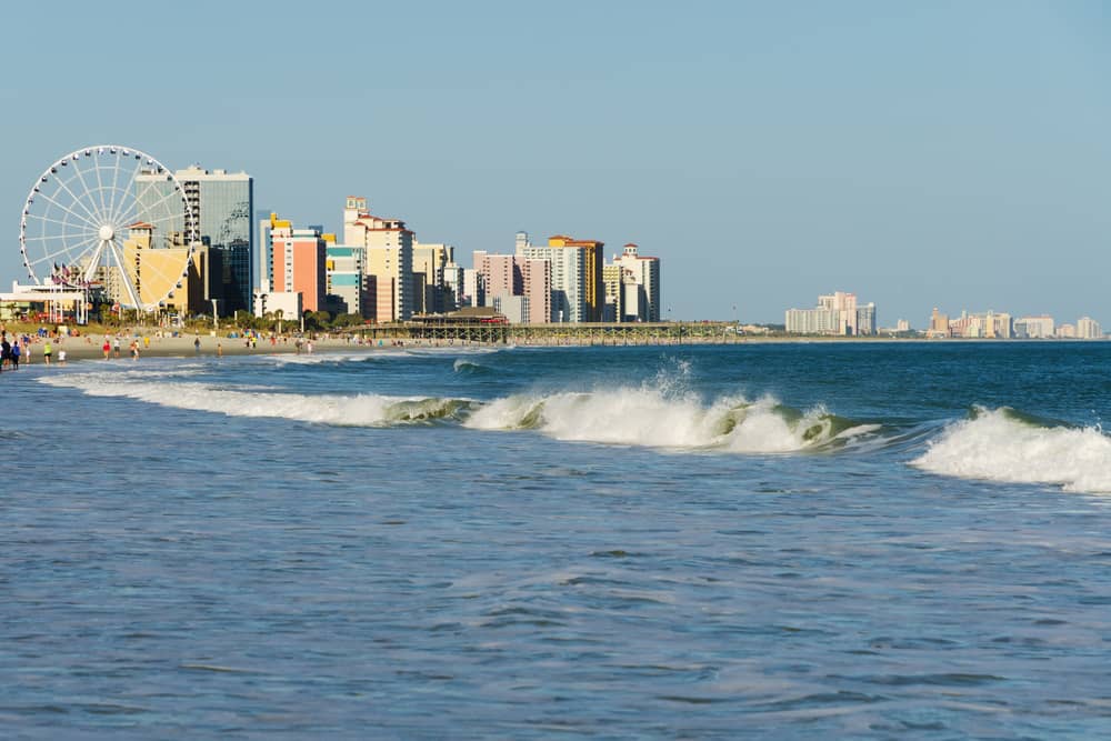 Beachfront cityscape with buildings, Ferris wheel, and ocean waves under a clear blue sky.
