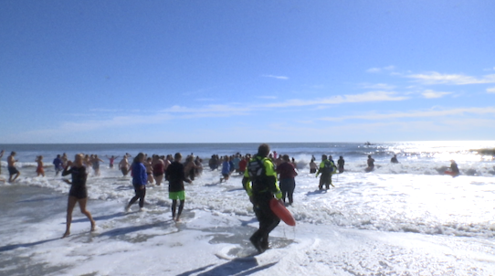 Crowd in swimwear braving cold beach water on a sunny day.