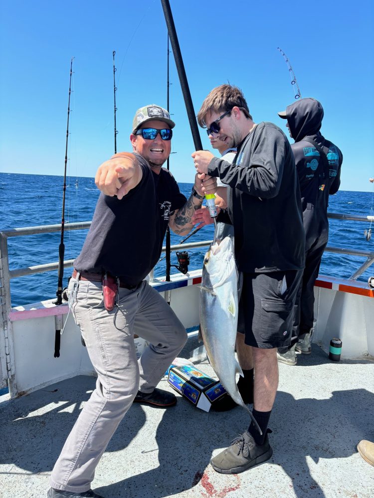 Group of people on a boat, one holding a large fish while another points excitedly, with a clear blue sky.