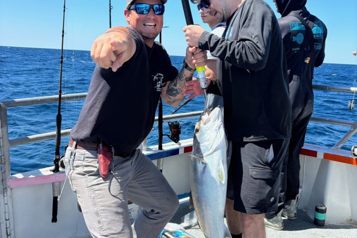 Group of people on a boat, one holding a large fish while another points excitedly, with a clear blue sky.
