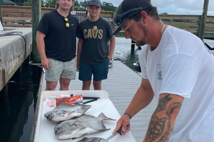 Three people on a dock; one is preparing fish on a table.
