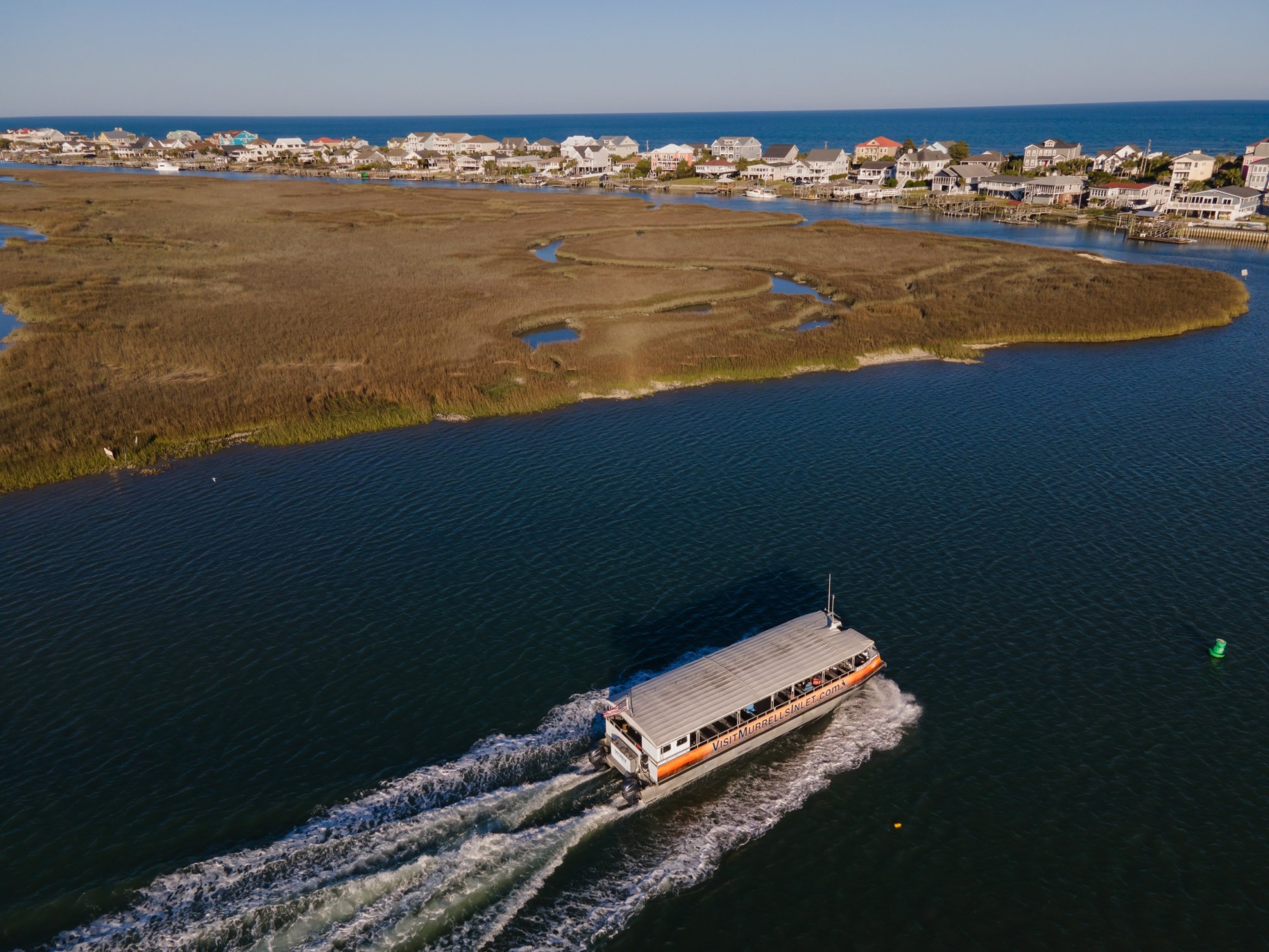 A boat travels through a waterway near marshland with houses in the background.