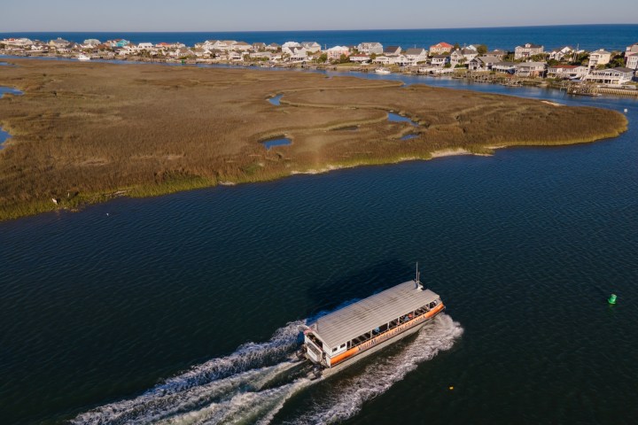A boat travels through a waterway near marshland with houses in the background.