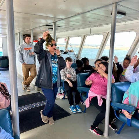 Person wearing sunglasses walks inside a boat holding a microphone, surrounded by seated passengers.