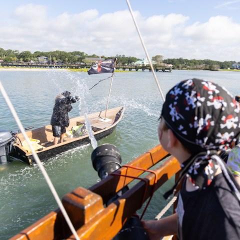 Children on a boat splash water at a small pirate-themed vessel in a river.