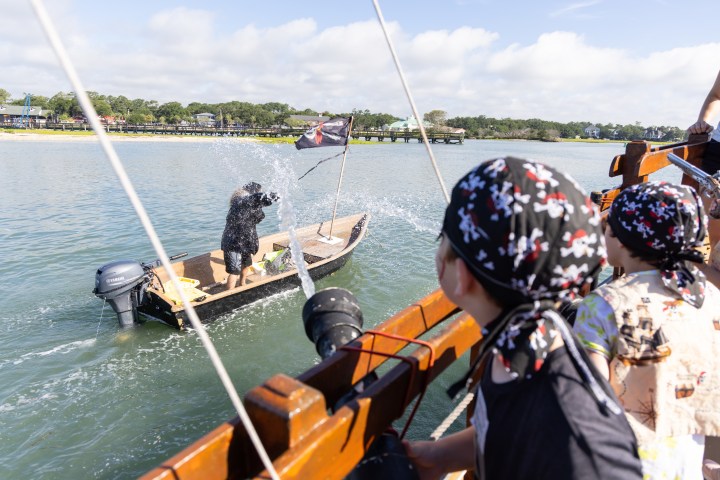 Children on a boat splash water at a small pirate-themed vessel in a river.