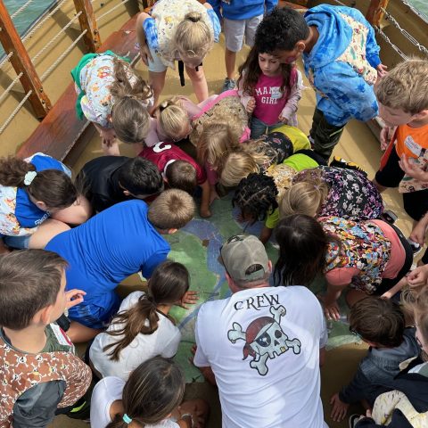 Group of children and adults leaning over and examining a large map on a wooden deck.