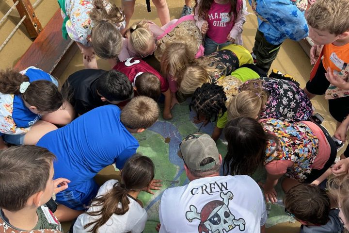 Group of children and adults leaning over and examining a large map on a wooden deck.