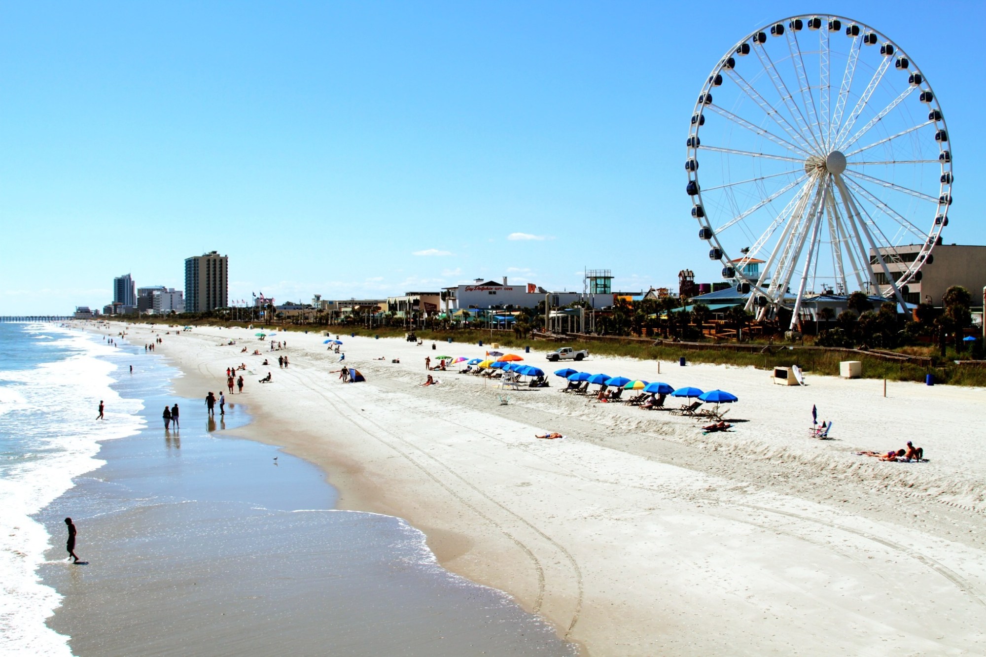 Beach scene with people, Ferris wheel, and umbrellas under clear blue sky.