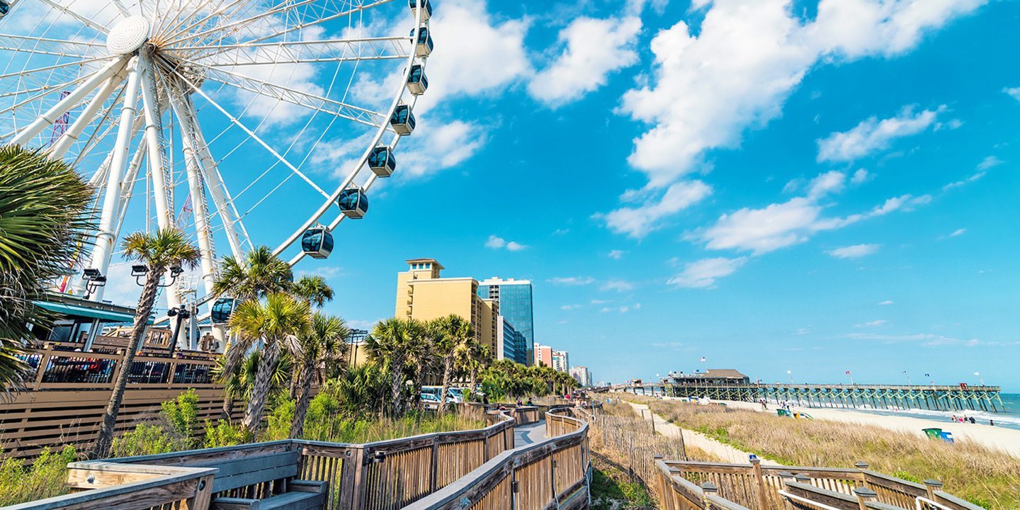 Ferris wheel near a boardwalk and beach with cityscape in the background under a blue sky.