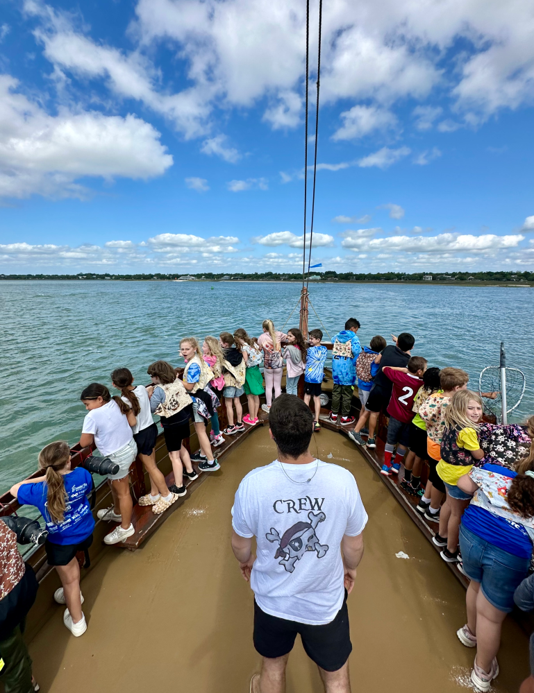 Group of children on a boat with adult wearing 'crew' shirt, overlooking water under a blue sky.